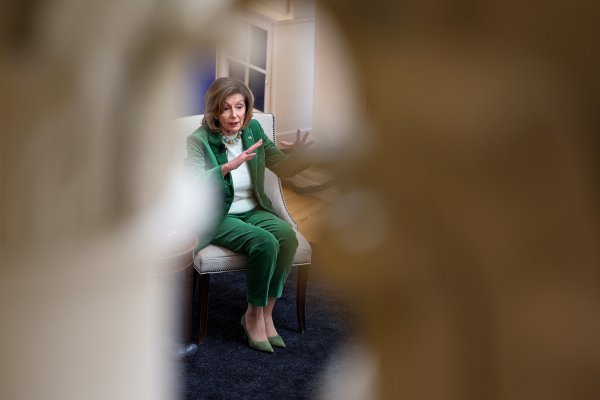 Speaker Emerita Nancy Pelosi sits on a chair, gesturing with both hands as she speaks, photographed through a blurred foreground that frames her.